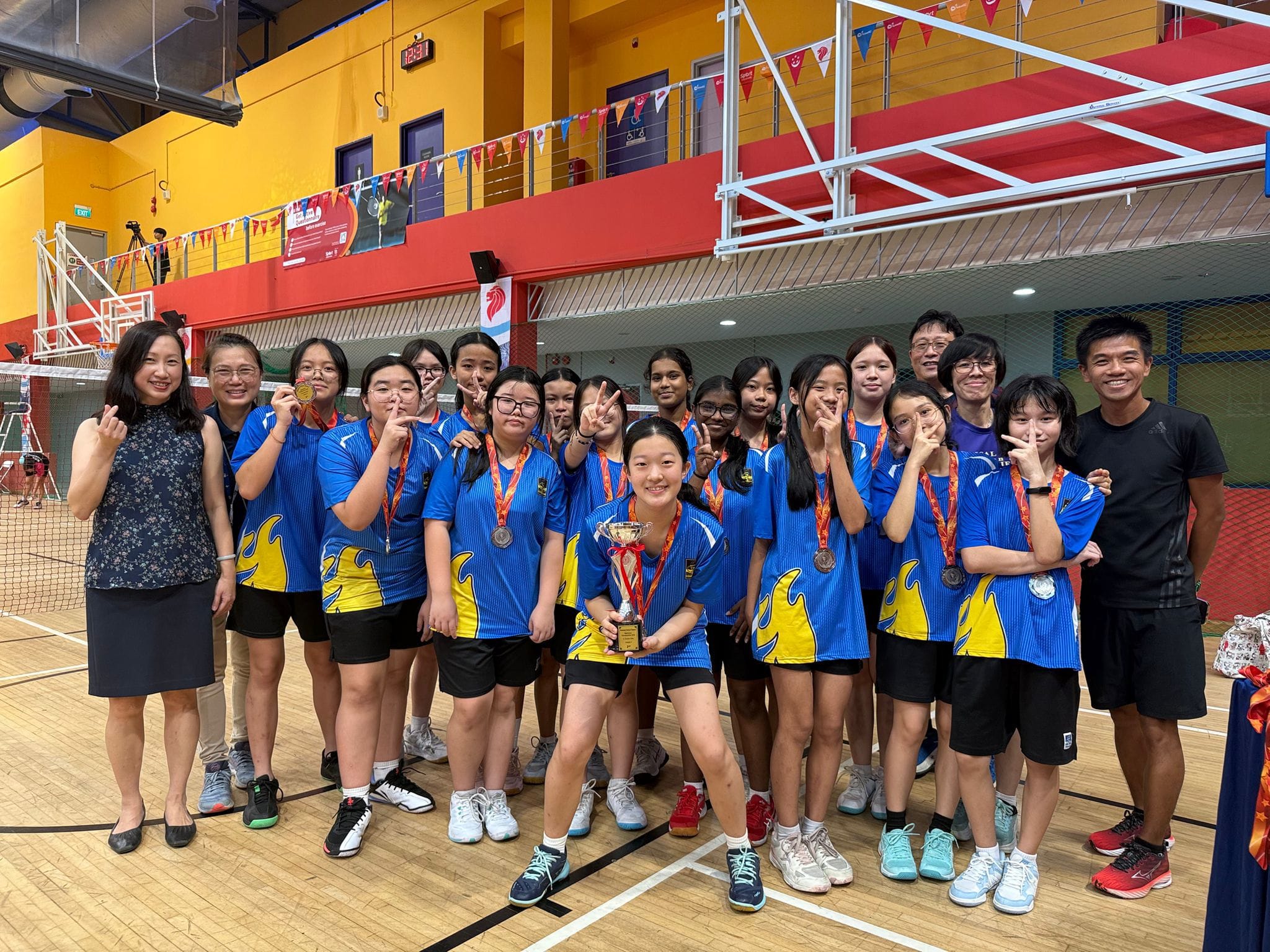 Photograph of a youth badminton team celebrating a victory in an indoor gymnasium, with players wearing matching blue and yellow uniforms and holding a trophy. Coaches and team members smile and pose under basketball hoops, with colorful banners and flags decorating the background. AI-generated content may be incorrect.